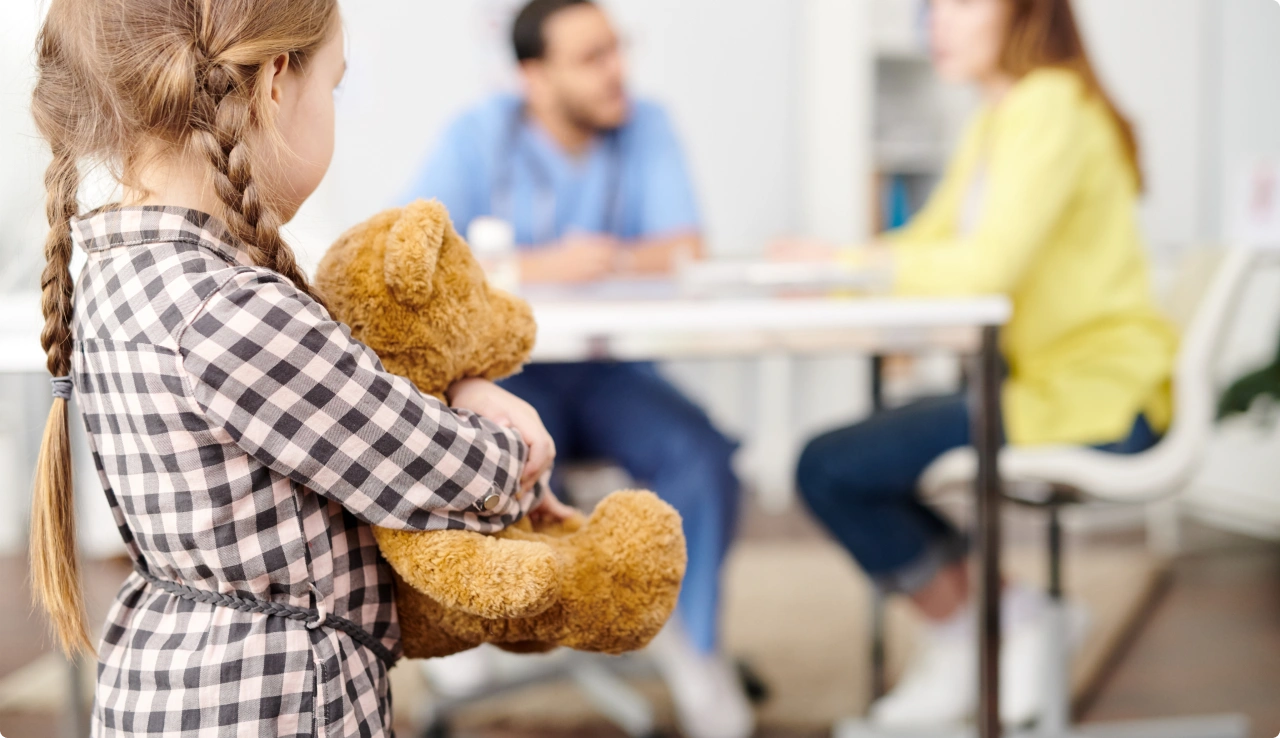 Niña con un oso de peluche mientras los adultos conversan al fondo
